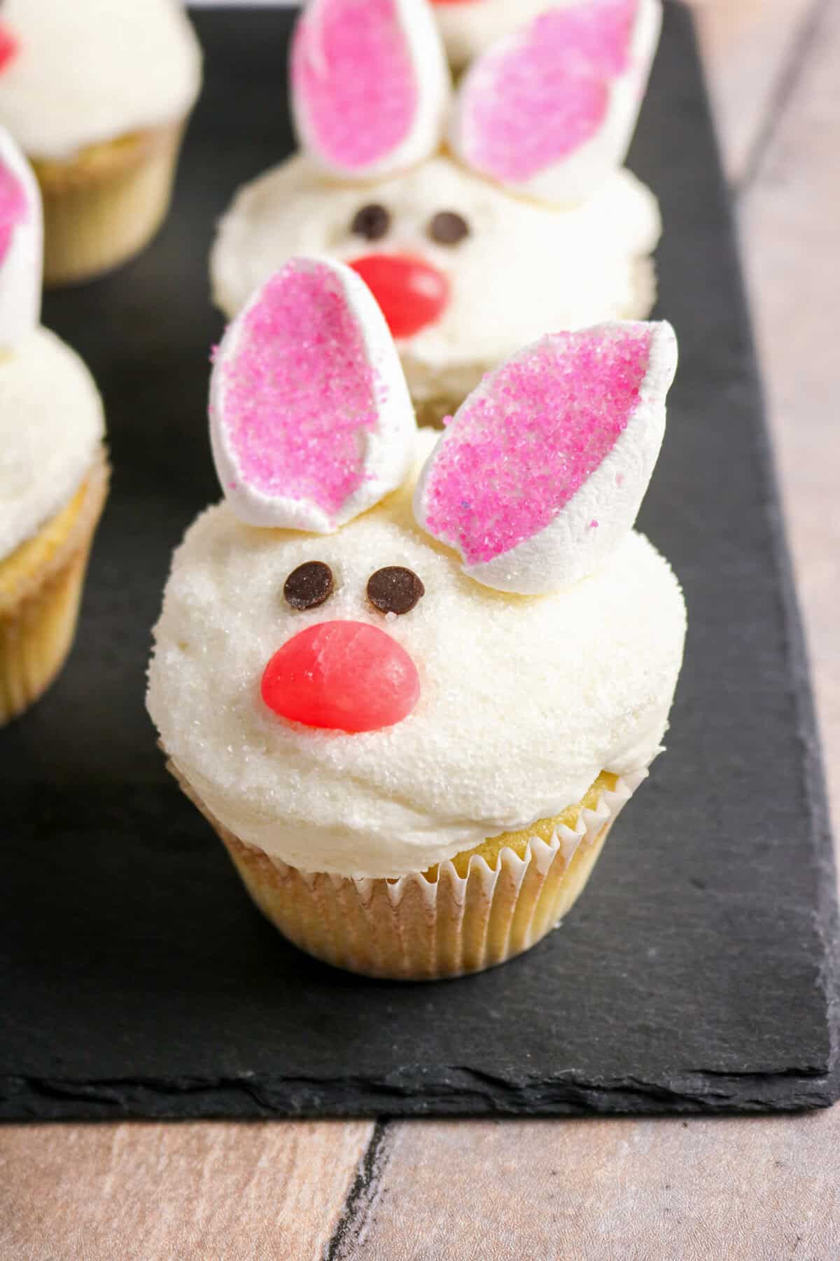 Decorated bunny cupcakes with marshmallow ears on a black slate serving board.
