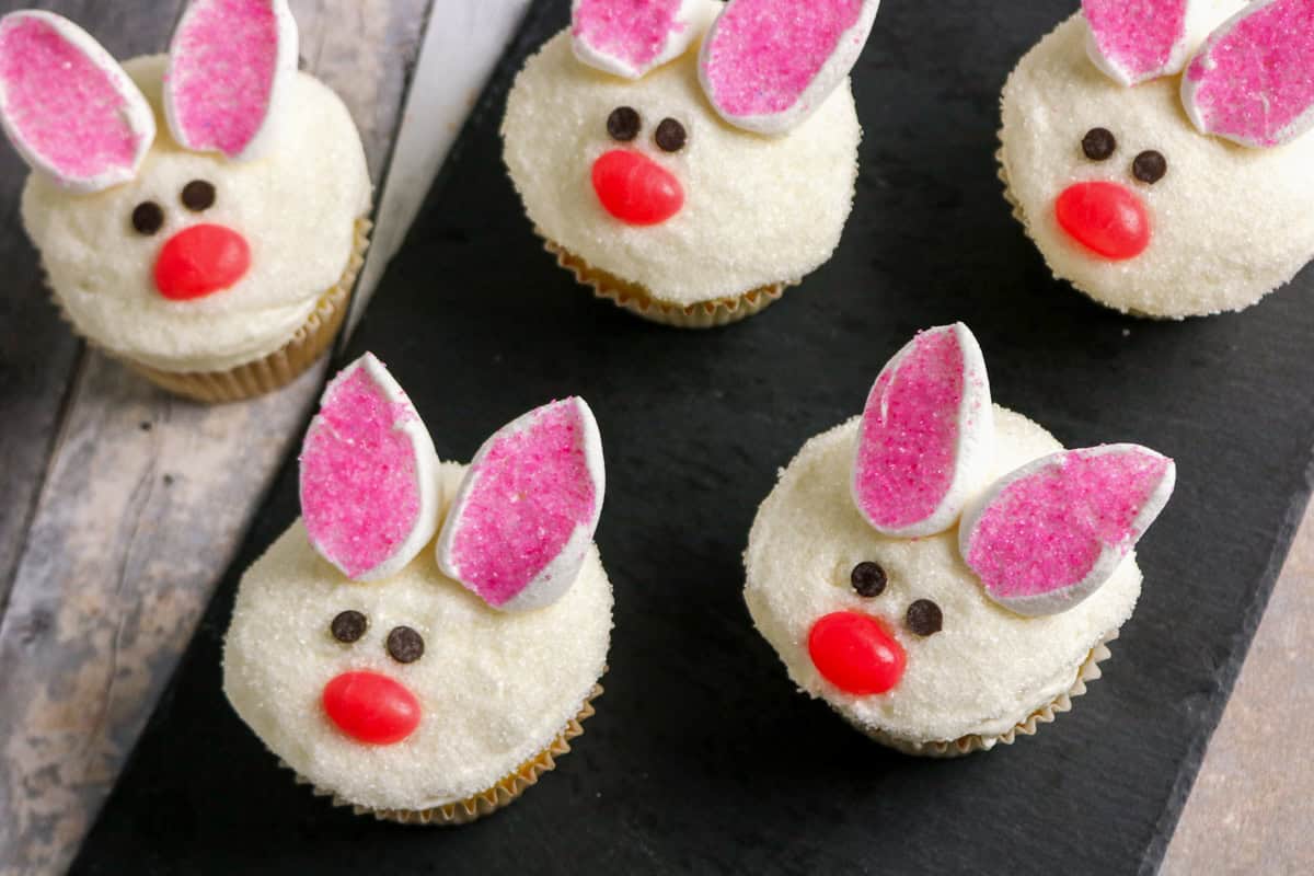 Five Easter bunny cupcakes decorated as bunny faces. Each cupcake has white sugar-coated frosting, marshmallow ears with pink sugar centers, mini chocolate chip eyes, and a red jelly bean nose.