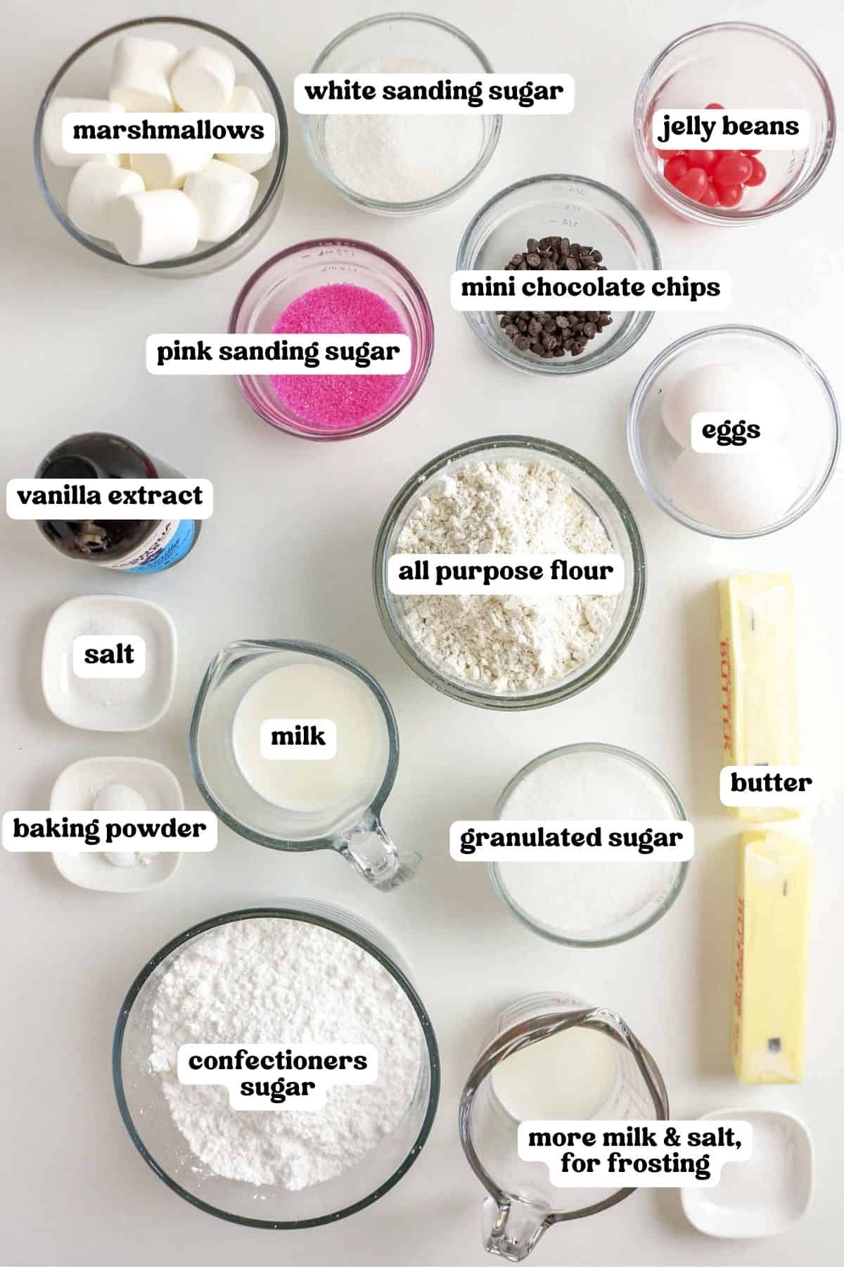 Baking ingredients arranged in bowls and labeled. Items include marshmallows, white sanding sugar, jelly beans, pink sanding sugar, mini chocolate chips, eggs, vanilla extract, salt, baking powder, milk, all-purpose flour, granulated sugar, butter, confectioners sugar, and more milk and salt for frosting.
