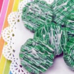 Candy-Dipped Green oreos with white chocolate drizzle on rainbow background.