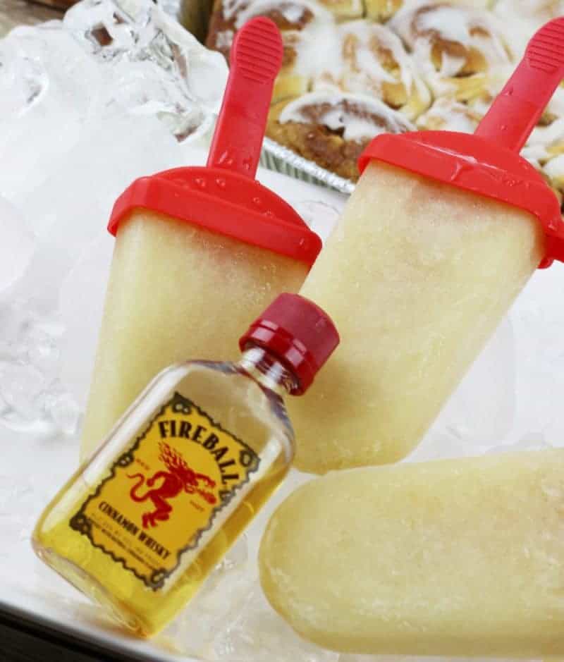 Fireball Whiskey Cinnamon Roll Ice Pops on tray of ice with cinnamon buns in the background and airplane-sized bottle of fireball whiskey infront of the ice pops.