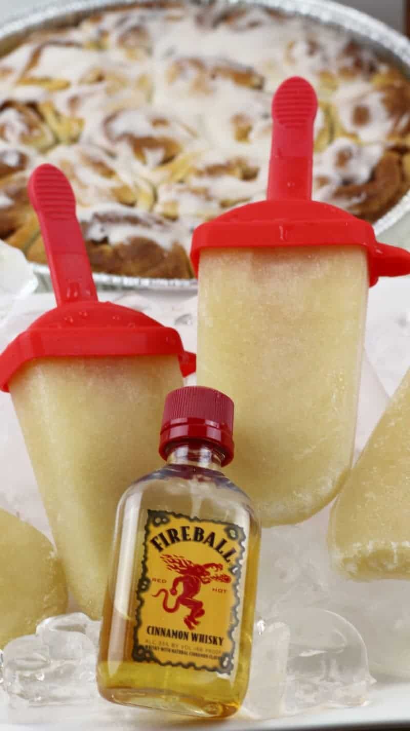 Fireball Whiskey cinnamon roll ice pops served on platter of ice with tray of cinnamon buns in the background and bottle of fireball cinnamon whiskey next to them