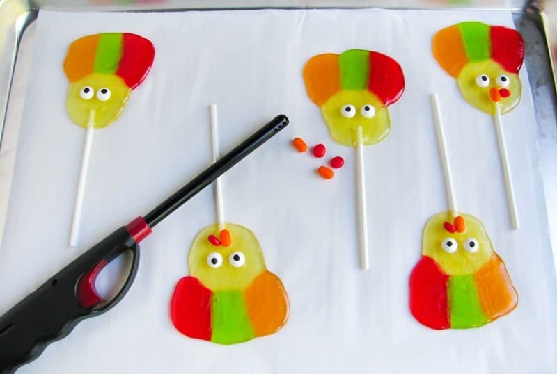 Handheld lighter, candies, and in process lollipops on lined baking sheet.