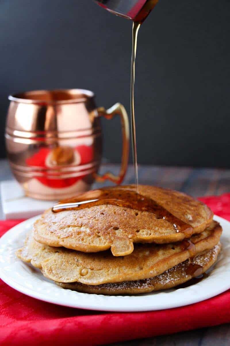 Maple syrup being drizzled over gingerbread pancakes stacked on a shite plate with bronze mug in background.