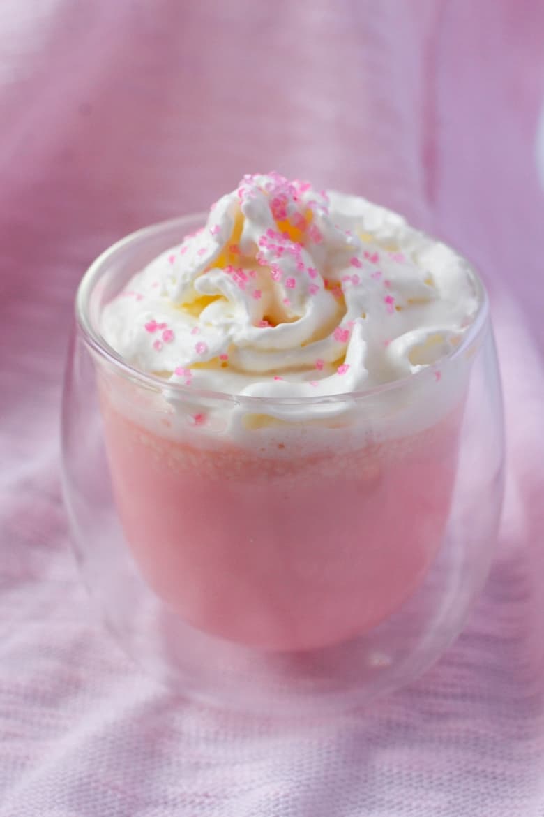 Pink velvet hot chocolate in a glass mug topped with whipped cream and pink sanding sugar.