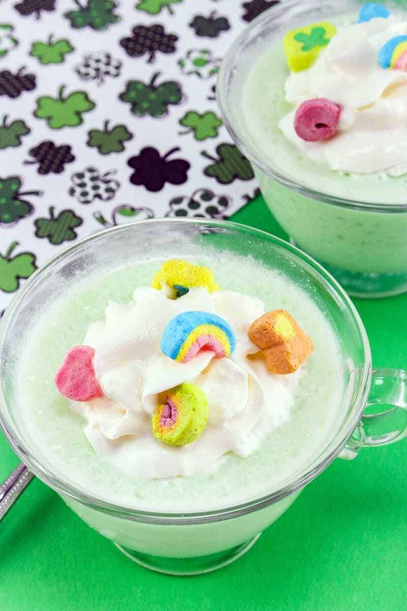 Frozen green drink with whipped cream and lucky charms marshmallows in a mug with shamrocks in the background.