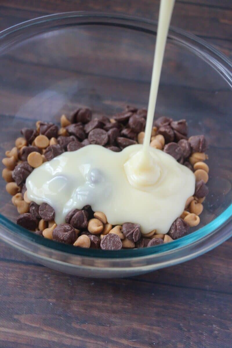 Peanut butter chips and chocolate chips with sweetened condensed milk being poured over them in a glass bowl.