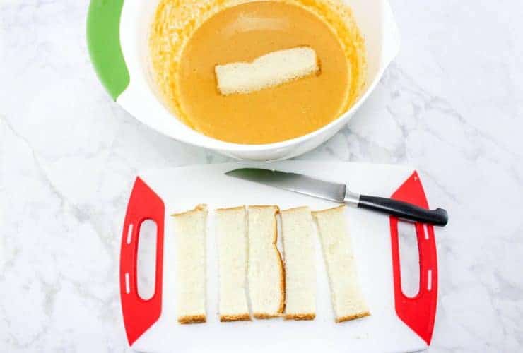 Bread and knife on cutting board. Bread is cut into thick strips and one strip is placed in pumpkin batter. 