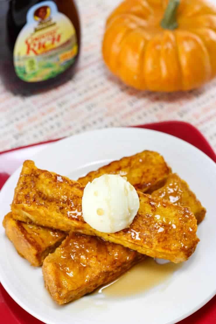Plate of french toast sticks with small pumpkin and bottle of pancake syrup in background.
