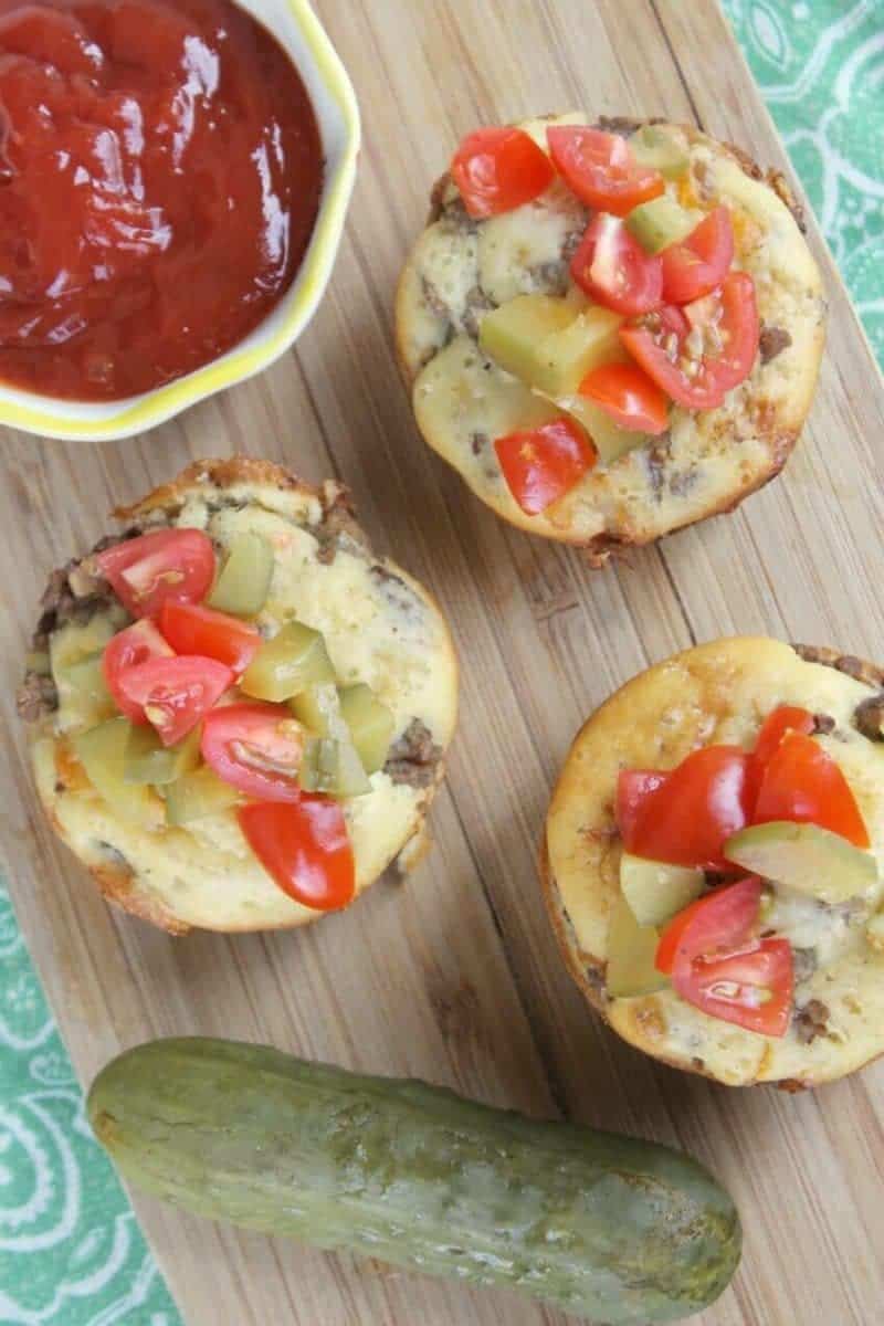 Cheeseburger muffins topped with chopped pickles and tomatoes on a wooden board with a bowl of ketchup and a pickle beside them.