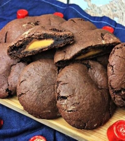 Rolo stuffed chocolate cookies piled on wooded cutting board with one cookie sliced in half to show rolo inside.