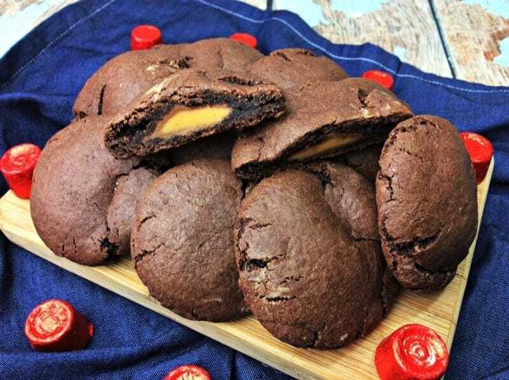 Rolo stuffed chocolate cookies piled on wooded cutting board with one cookie sliced in half to show rolo inside.