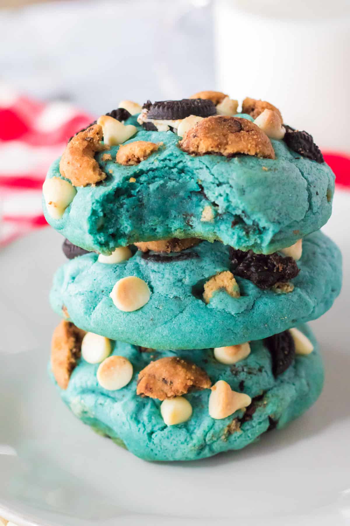 Thick blue cookie monster cookies stacked on top of one another with a bite taken out of the top cookies and a glass of milk in the background.
