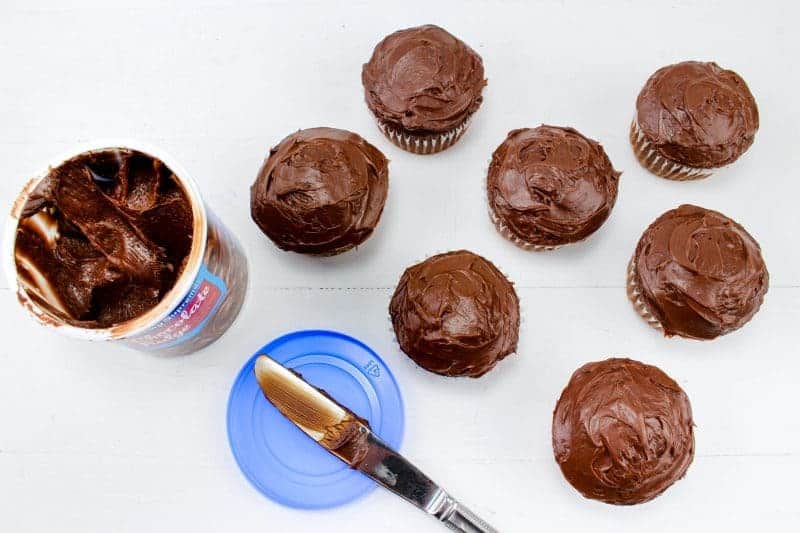 Seven chocolate cupcakes with chocolate frosting next to tub of chocolate frosting and butter knife.