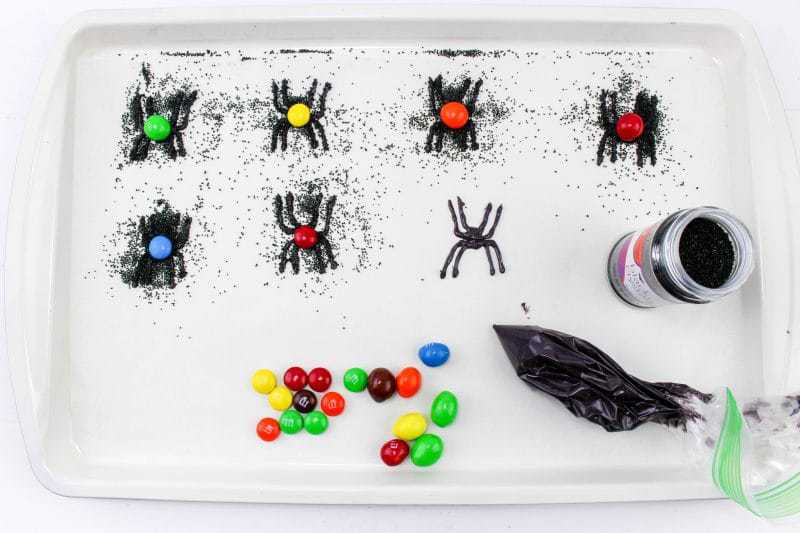 Candy spiders covered with black sanding sugar on a lined baking sheet. Some have colorful M&Ms on their backs.