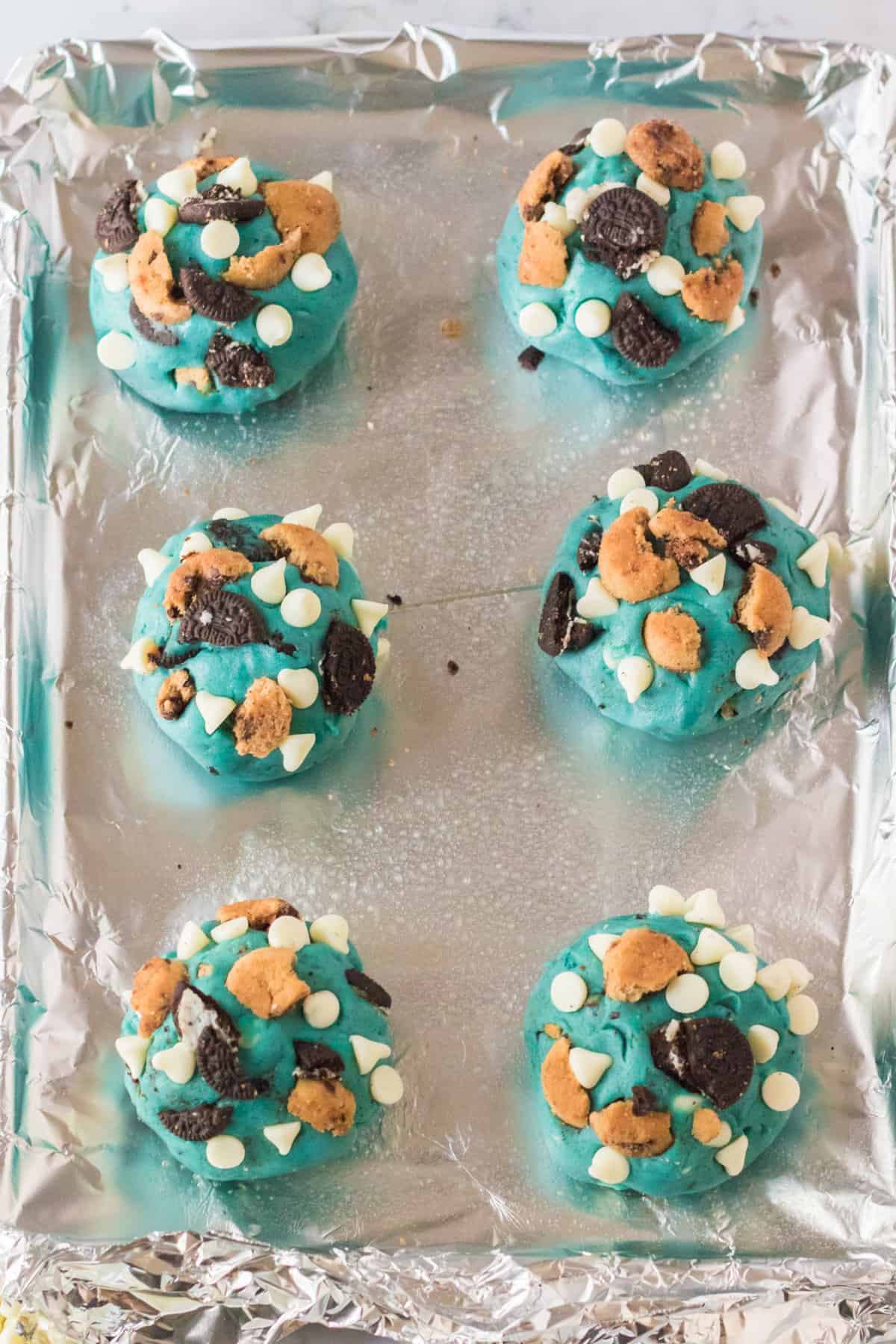 Large balls of blue cookie dough topped with crushed cookies and white chocolate chips. 