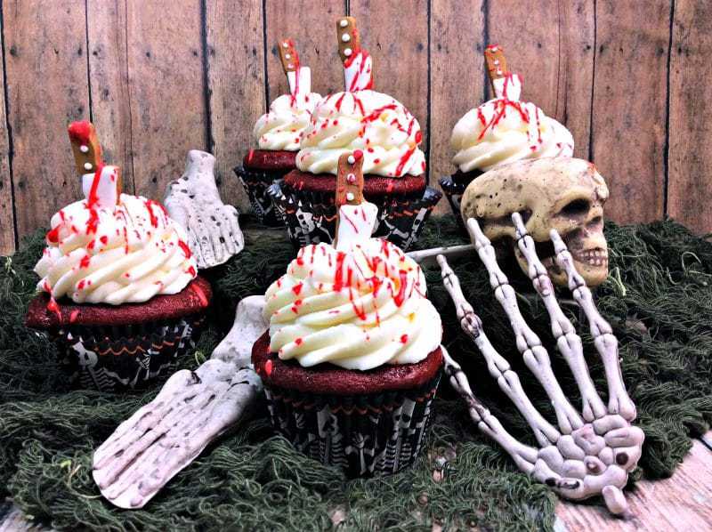 Red velvet cupcakes topped with cream cheese frosting, knives, and edible blood splatter. Bones and skull decorations are around the cupcakes.