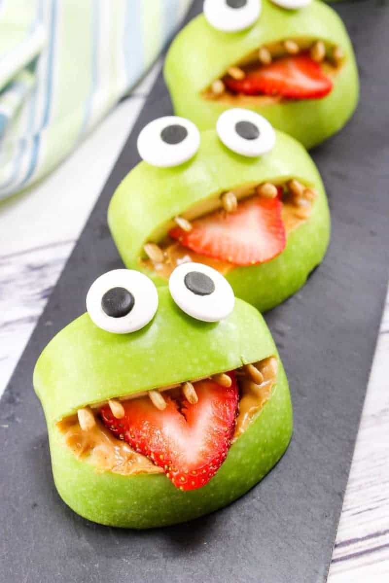 Kids snack made with sliced green apples, and a peanut butter mouth, sunflower seed teeth, strawberry tongues and candy eyes.