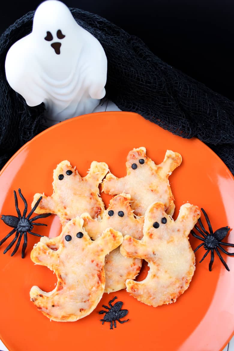 Ghost shaped pizzas with olive eyes served on an orange plate surrounded by Halloween decorations.