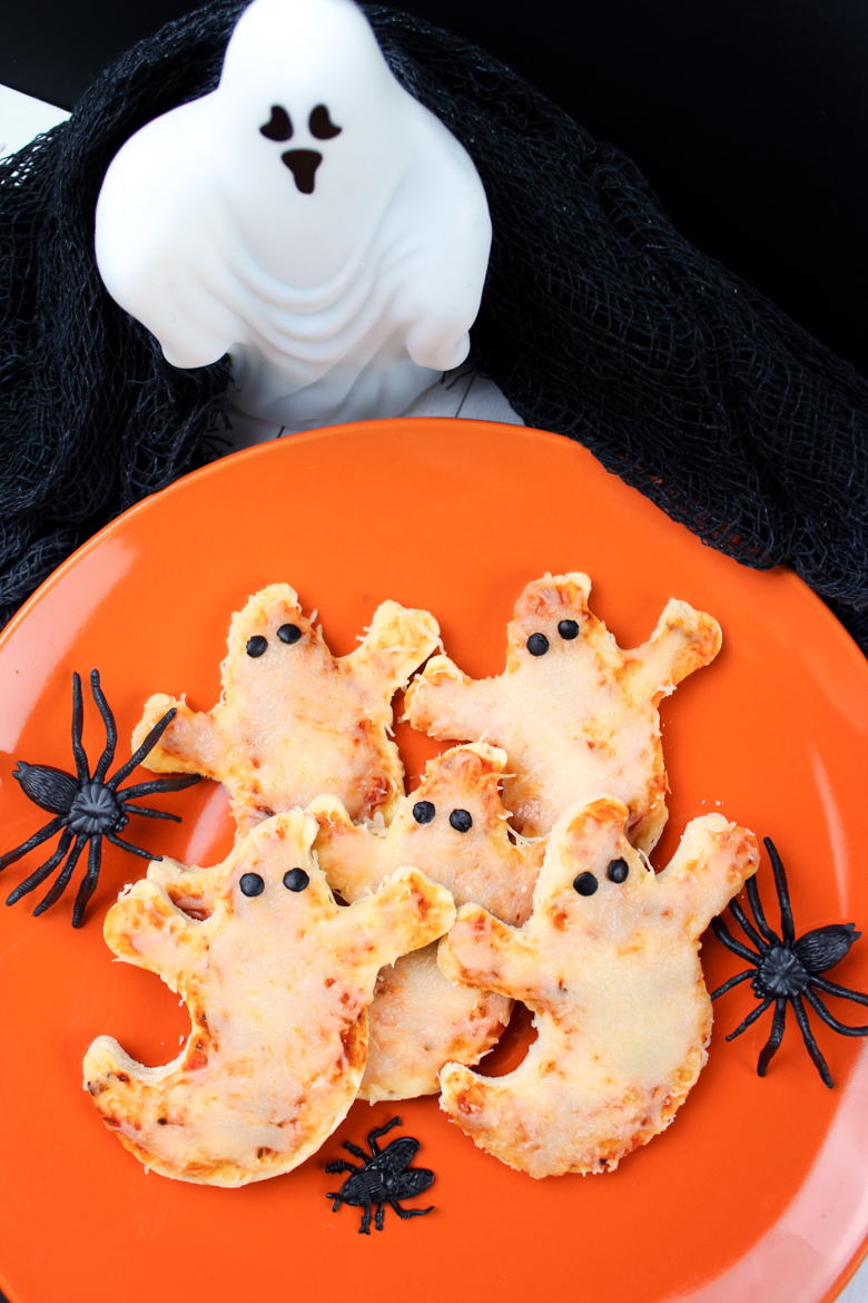Ghost shaped pizzas with olive eyes served on an orange plate surrounded by Halloween decorations.