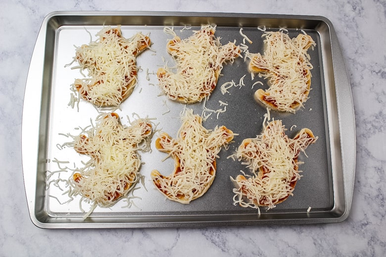 6 ghost pizza crusts covered in pizza sauce and shredded mozzarella cheese on a baking sheet.
