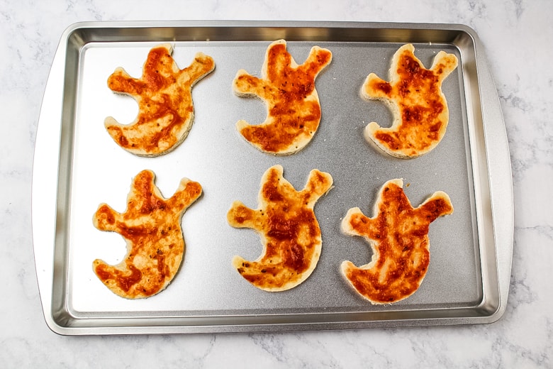6 ghost pizza crusts covered in pizza sauce on a baking sheet.