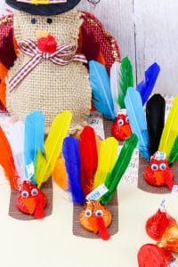 A group of small Thanksgiving-themed turkey crafts made using Hershey’s Kisses as the body, with colorful feathers attached at the back, googly eyes, and orange beaks. Each sits on a small brown base. A burlap turkey decoration is placed in the background.