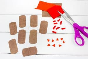 Brown paper bases, small orange felt triangles and wattles, and a pair of purple-handled scissors are laid out on a white surface for assembling Hershey’s Kiss turkey crafts.