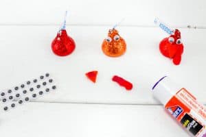 Three Hershey’s Kisses on a white surface: one plain, one with googly eyes, and one with eyes and a red felt wattle. Nearby are small googly eyes, orange felt triangles for beaks, red wattles, and a glue stick.