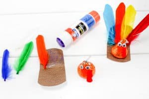 Crafting in progress: a glue stick, cut feathers, a Hershey’s Kiss with eyes and beak, and a brown paper base with a feather attached. A finished turkey craft is shown as a reference.