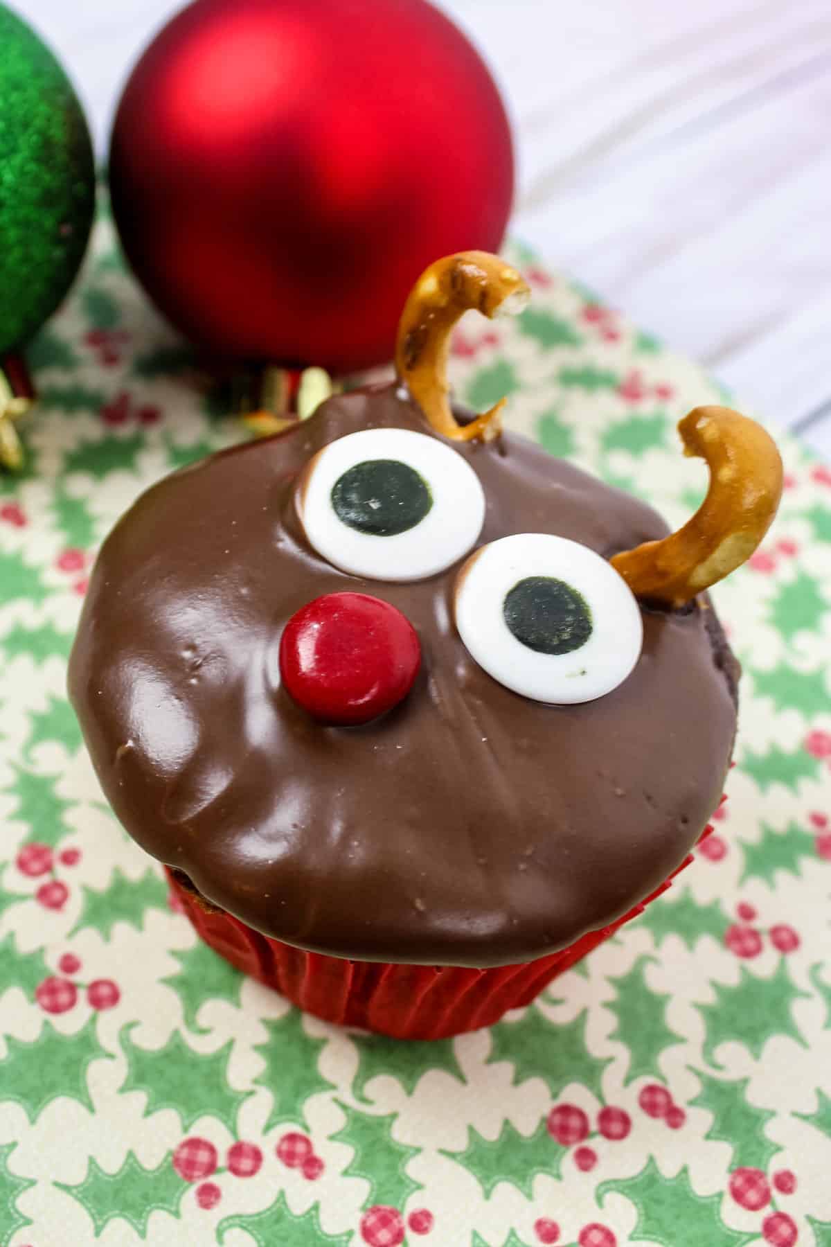 Chocolate cupcake with chocolate icing decorated with pretzel antlers, candy eyes, and a red m&m, to look like Rudolph the red nose reindeer.