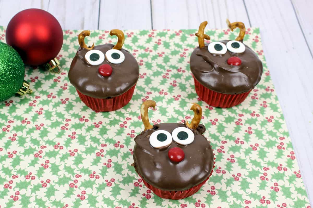 Three chocolate reindeer cupcakes on Christmas printed napkin with ornaments beside them.