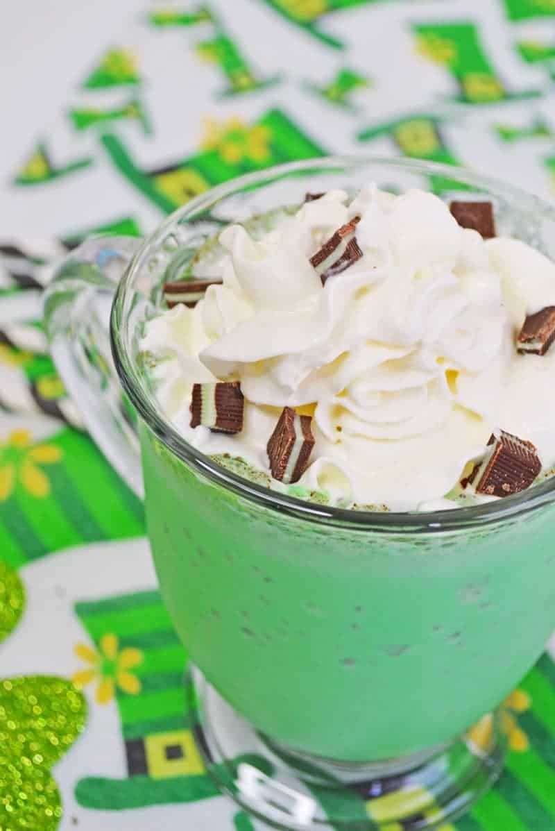 Reads: Shamrock Shake. Green mint chocolate chip milkshake in glass mug with whipped cream, chopped Andes Mints, and a white and black striped paper straw. The mug sits on a leprechaun hat tablecloth. 