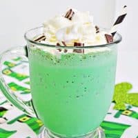 Bright green mint chocolate chip milkshake in glass mug with whipped cream, chopped Andes Mints, and a white and black striped paper straw. The mug sits on a leprechaun hat tablecloth.