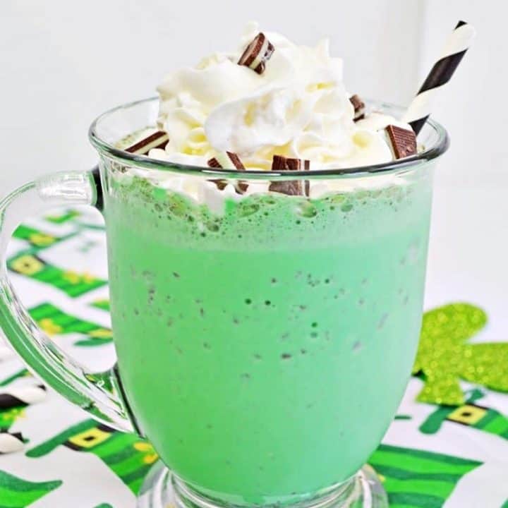 Bright green mint chocolate chip milkshake in glass mug with whipped cream, chopped Andes Mints, and a white and black striped paper straw. The mug sits on a leprechaun hat tablecloth.
