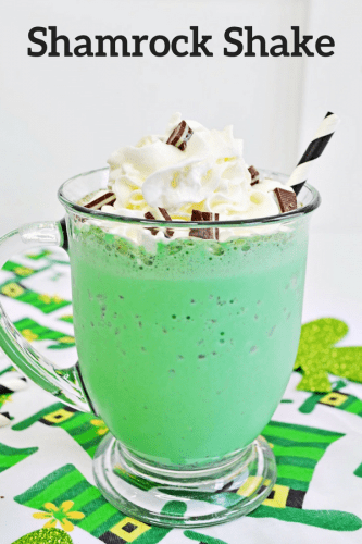 Reads: Shamrock Shake. Green mint chocolate chip milkshake in glass mug with whipped cream, chopped Andes Mints, and a white and black striped paper straw. The mug sits on a leprechaun hat tablecloth.