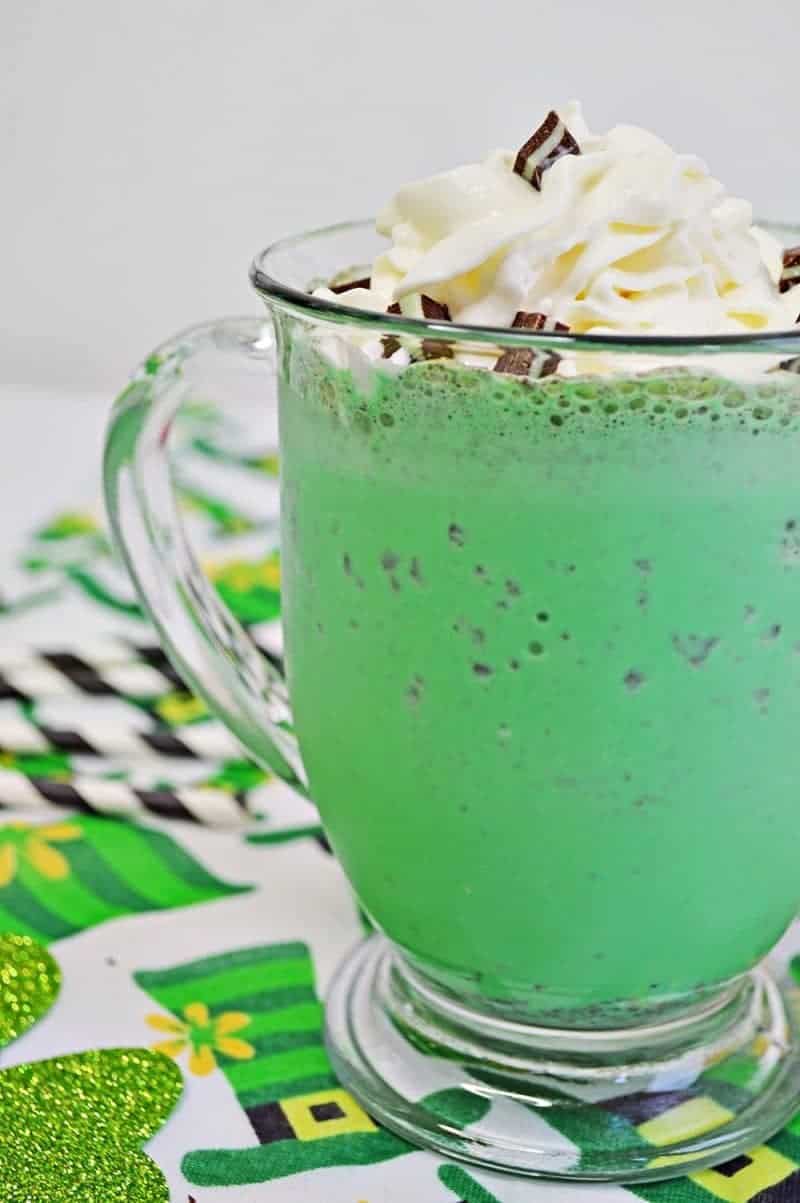 Bright green mint chocolate chip milkshake in glass mug with whipped cream, chopped Andes Mints, and a white and black striped paper straw. The mug sits on a leprechaun hat tablecloth. 