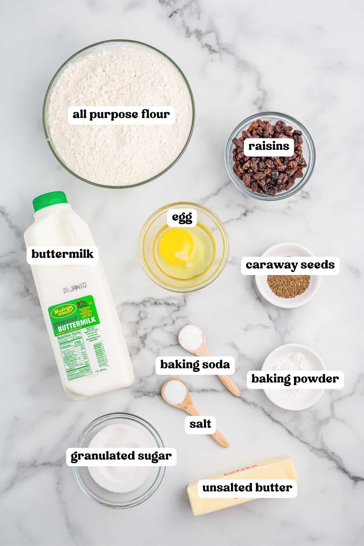 Overhead view of Irish soda bread ingredients: including all-purpose flour, raisins, caraway seeds, egg, buttermilk, granulated sugar, baking soda, baking powder, salt, and unsalted butter.
