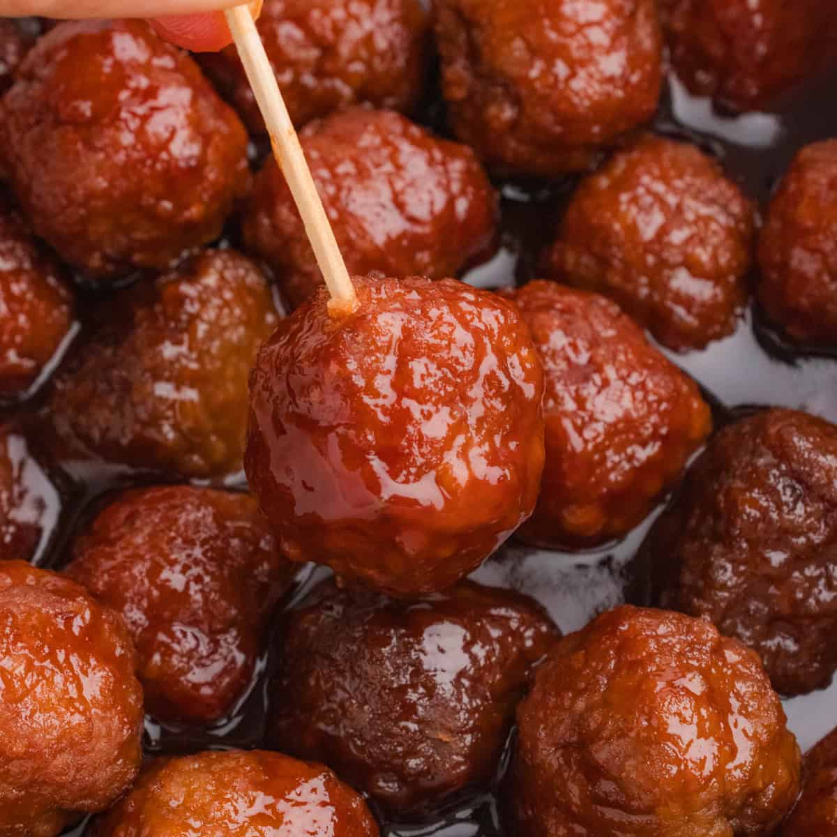 A close-up of a glazed meatball covered in thick barbecue and grape jelly sauce, held by a wooden toothpick. More crockpot meatballs are visible in the background, coated in the glossy sauce.