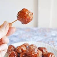 Crockpot grape jelly and BBQ sauce meatballs served on a plate with a hand reaching in to lift a single meatball with a toothpick.