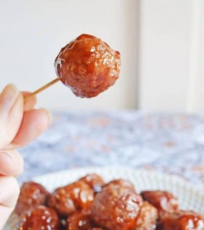 Crockpot grape jelly and BBQ sauce meatballs served on a plate with a hand reaching in to lift a single meatball with a toothpick.