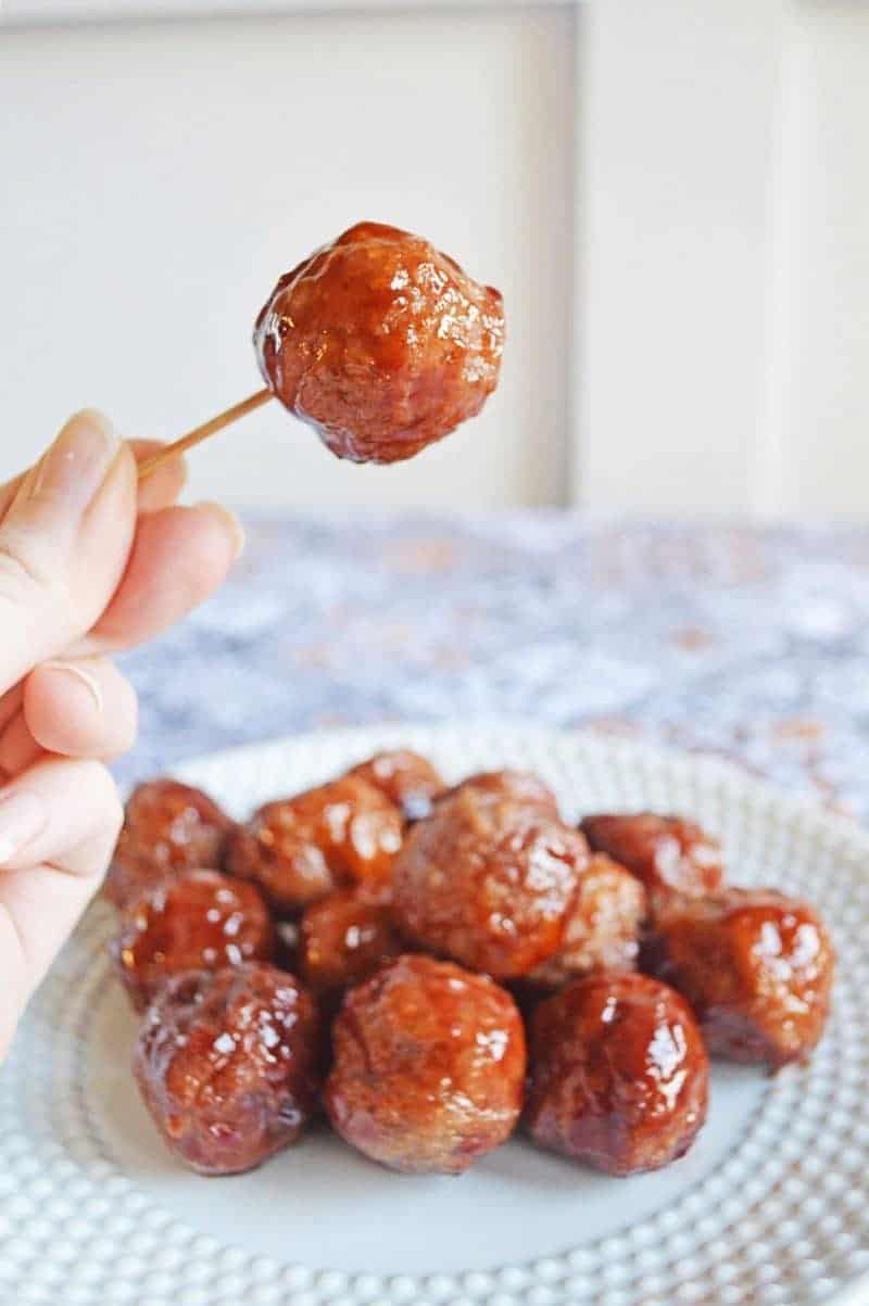 Meatballs with grape jelly and bbq sauce on a plate with a single meatball picked up with a hand holding a tooth pick.