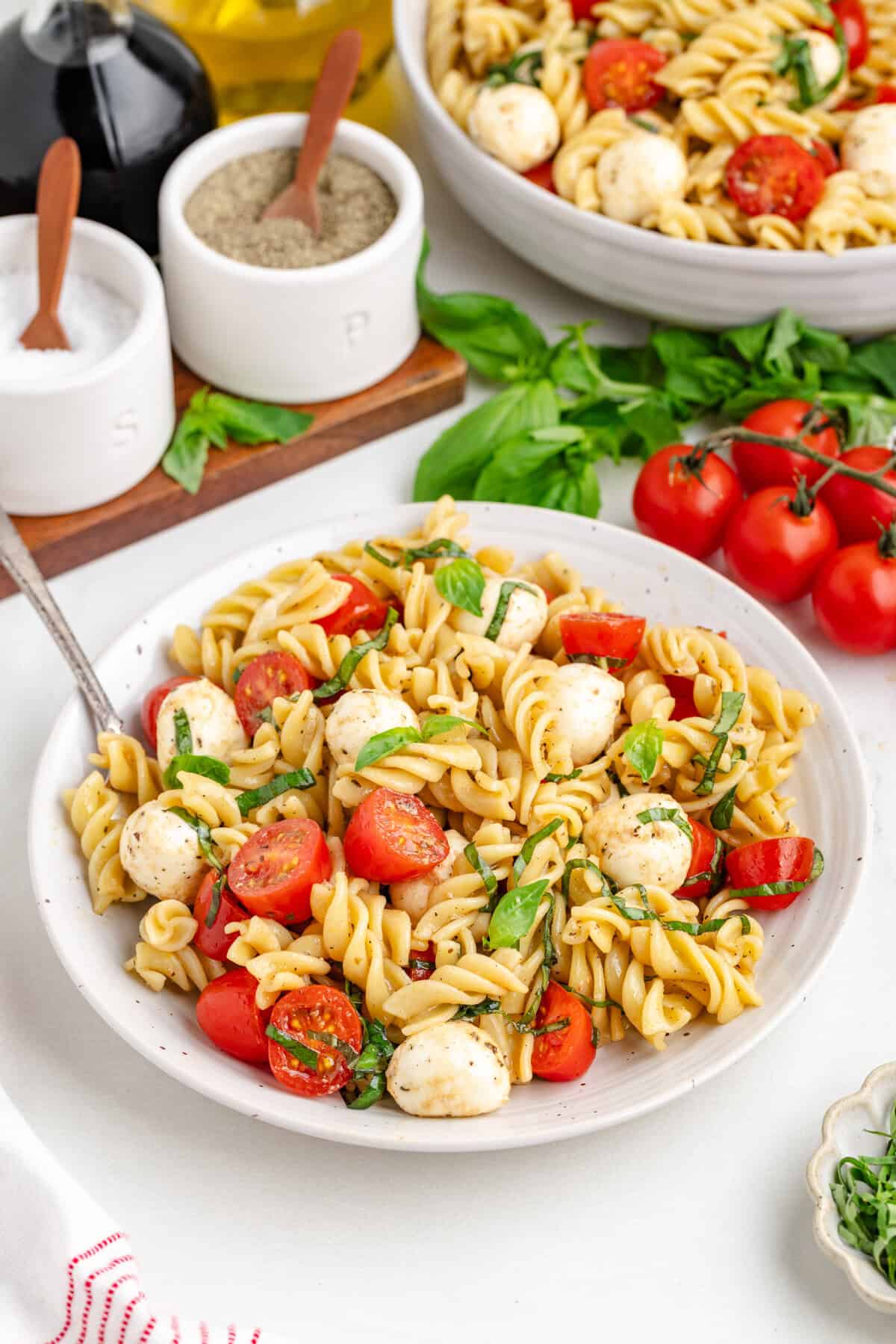 Pasta salad with basil, tomatoes and mozzarella, with ingredients in the background.