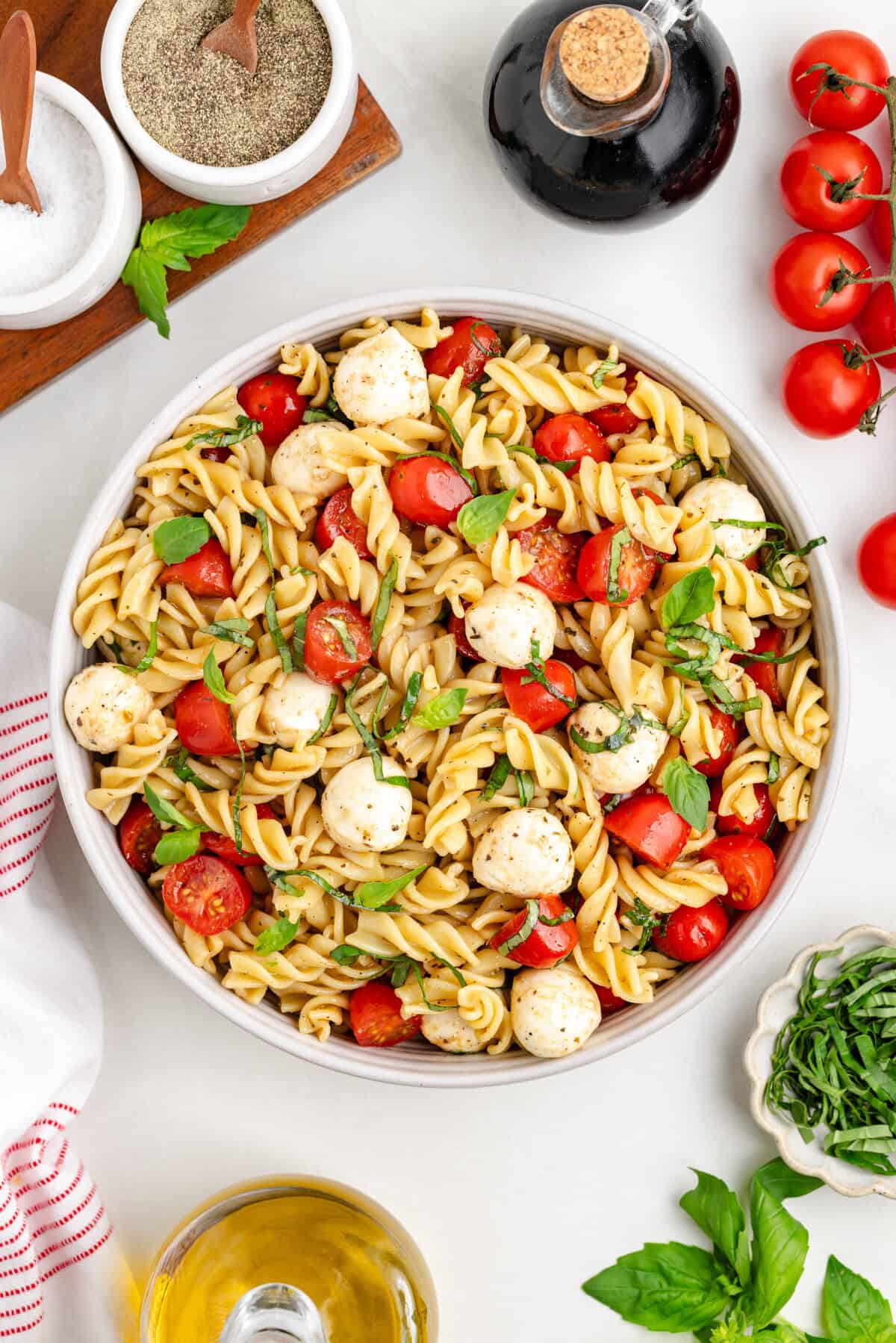Overhead view of a large bowl of caprese pasta salad with rotini, cherry tomatoes, mozzarella balls, and basil. Around the bowl are a bottle of balsamic vinegar, salt, pepper, olive oil, fresh basil leaves, and cherry tomatoes on the vine.