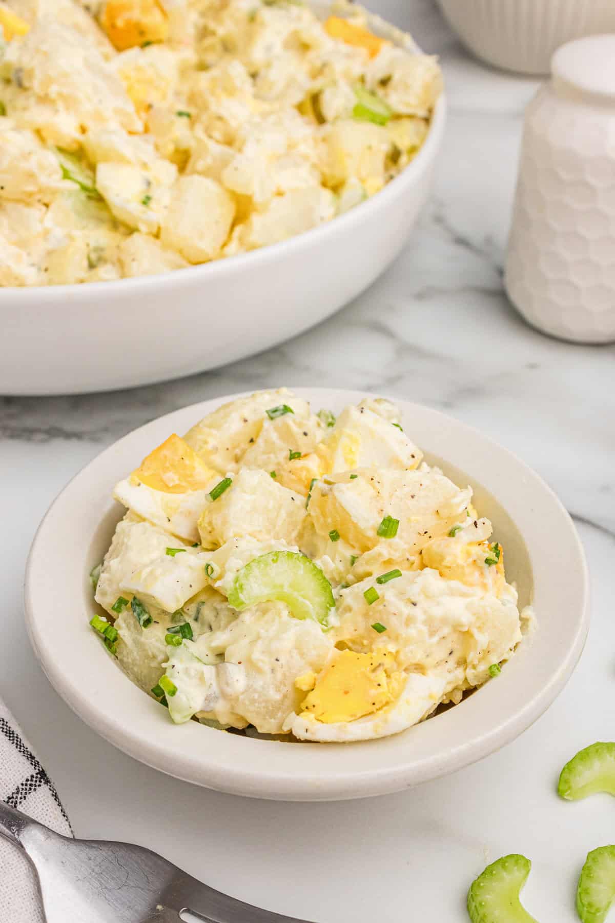 A small white bowl filled with a serving of potato salad placed in front of a larger bowl of the same dish. The salad includes diced potatoes, celery, eggs, and herbs in a creamy dressing.