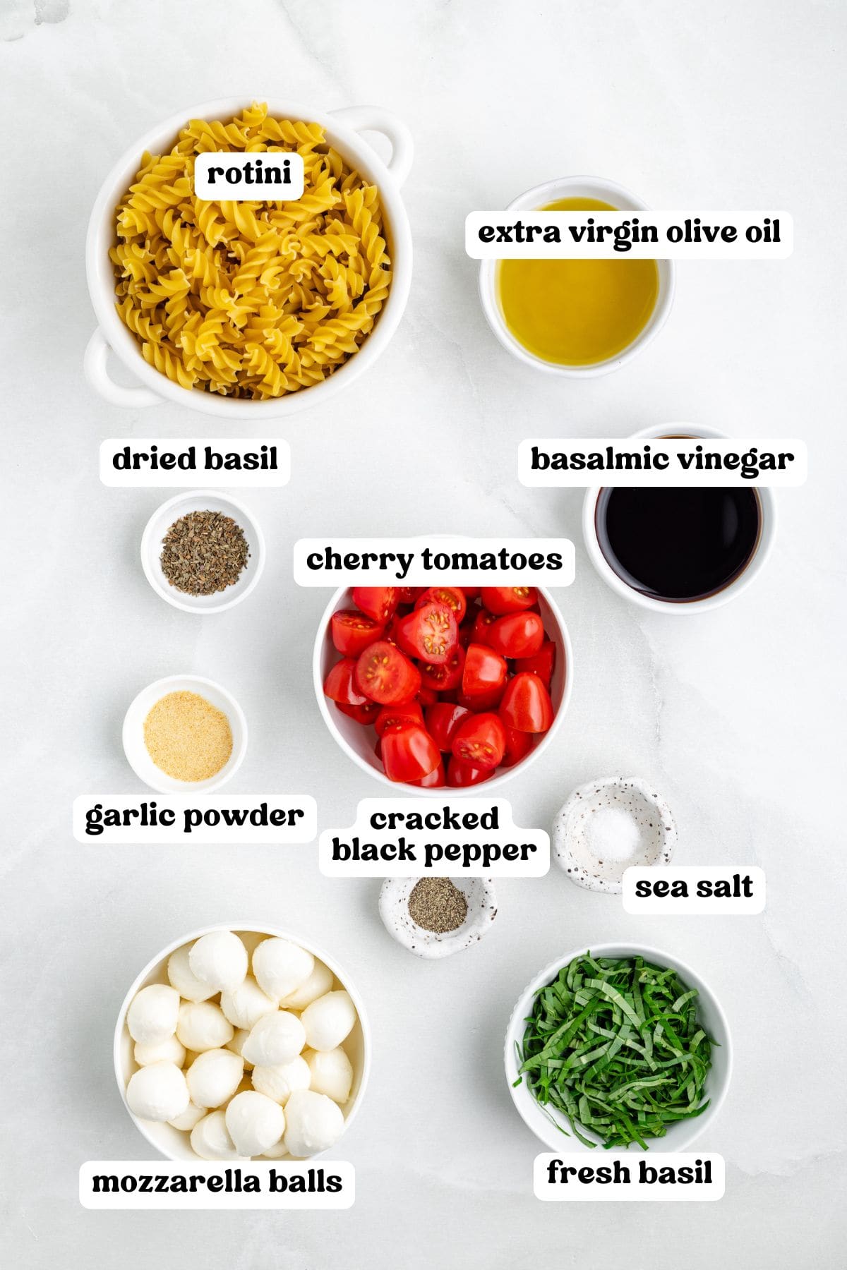 Overhead view of labeled ingredients in bowls for making Caprese pasta salad. Labels include rotini, extra virgin olive oil, balsamic vinegar, dried basil, garlic powder, cherry tomatoes, cracked black pepper, sea salt, mozzarella balls, and fresh basil.