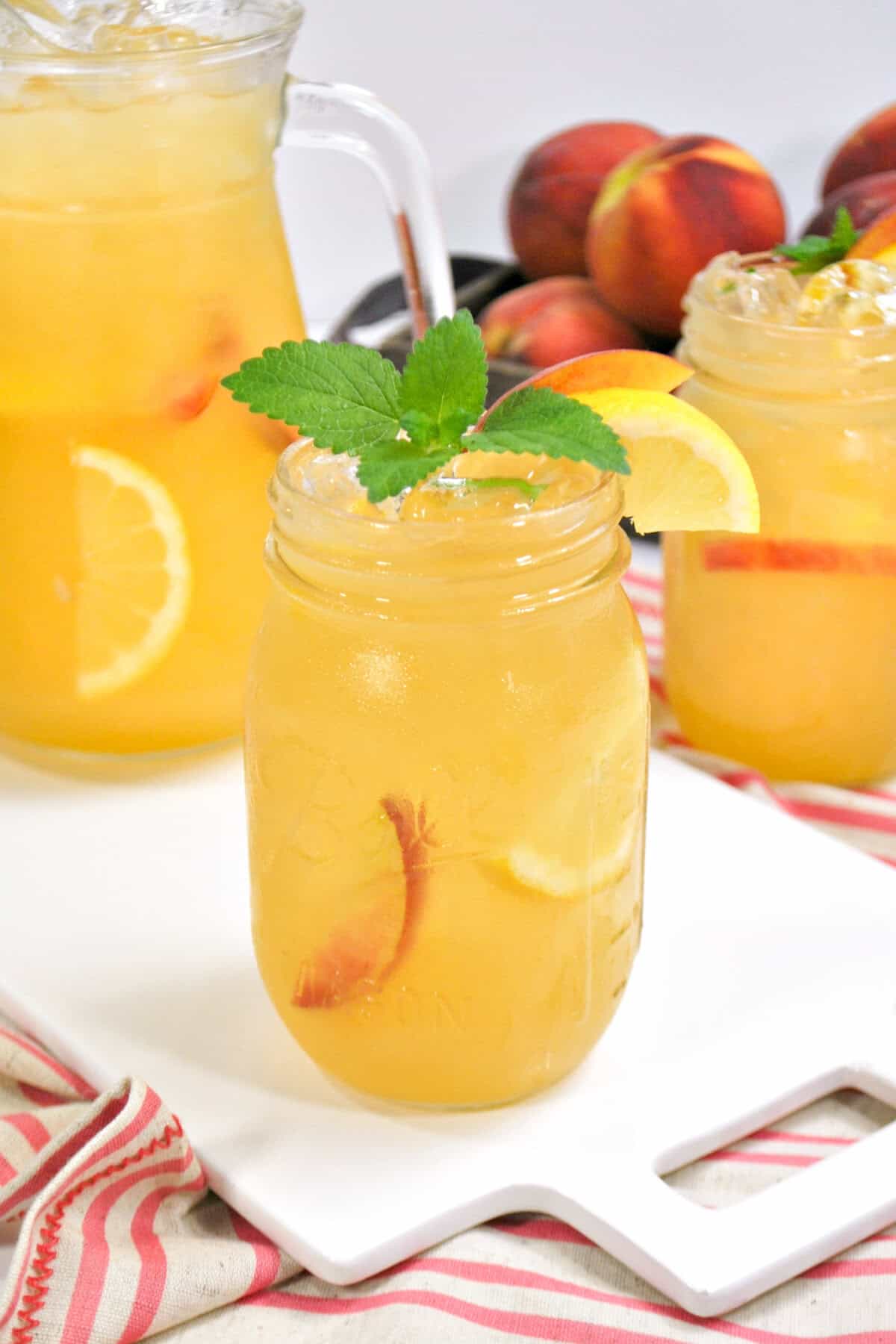 A mason jar of peach lemonade garnished with lemon, peach slice, and mint, with a pitcher and another jar and fresh peaches in the background.