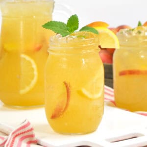 A mason jar of peach lemonade with lemon, peach slices, and mint, set in front of a pitcher and another jar.