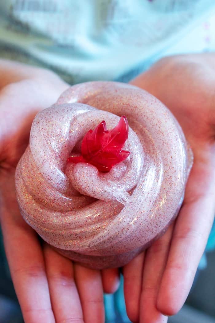 A cinnamon-scented fall slime with glitter in a child's hands with a small red plastic leaf toy on top of it.