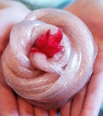 A cinnamon-scented fall slime with glitter in a child's hands with a small red plastic leaf toy on top of it.